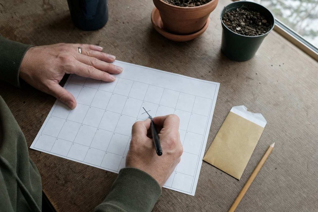 hands marking a date on a plain paper wall calendar beside a packet of seeds