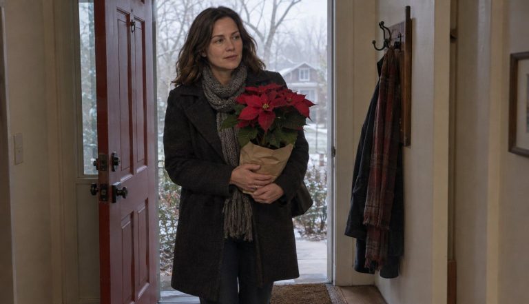 a woman in a winter coat carrying a paper-wrapped poinsettia through her front door